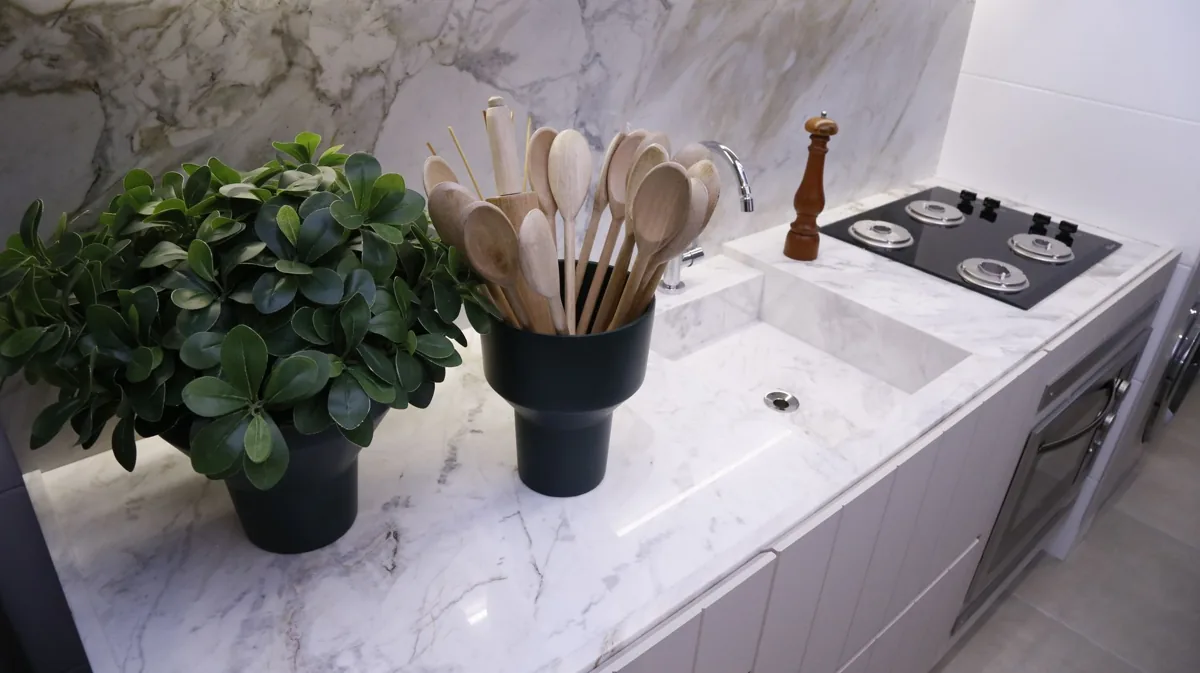 Sleek minimalist kitchen with marble counter, wooden spoons, potted plant, modern fixtures, and stovetop.